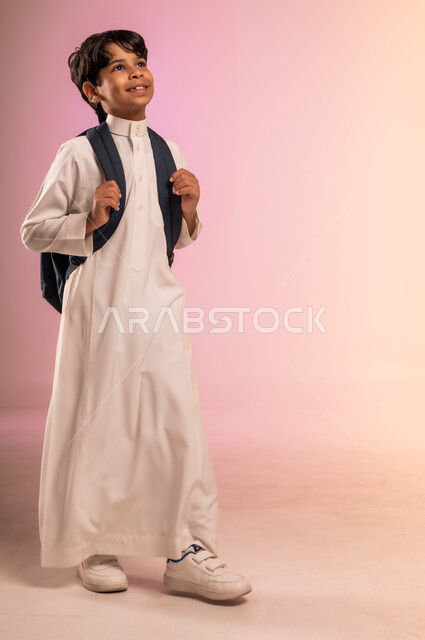 Passion and striving to achieve the dream of success and excellence, portrait of a Saudi Arabian Gulf student wearing traditional dress carrying a school backpack looking up with gestures of hope and cheerfulness, back to school season in Saudi Arabia, full body image, colorful background