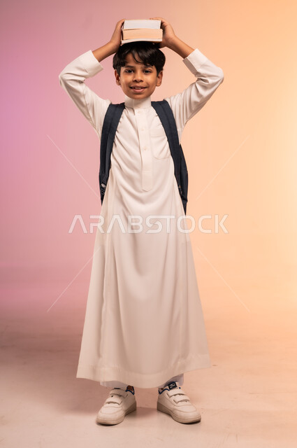  النظر للكاميرا بإيماءات السرور، الاستعداد للذهاب إلى المدرسة، بورتريه لطالب عربي خليجي سعودي يرتدي الثوب التقليدي يحمل حقيبة الظهر المدرسية يضع الكتب العلمية فوق رأسه، موسم العودة للمدارس في المملكة العربية السعودية، صورة الجسم بالطول الكامل، خلفية ملونة
