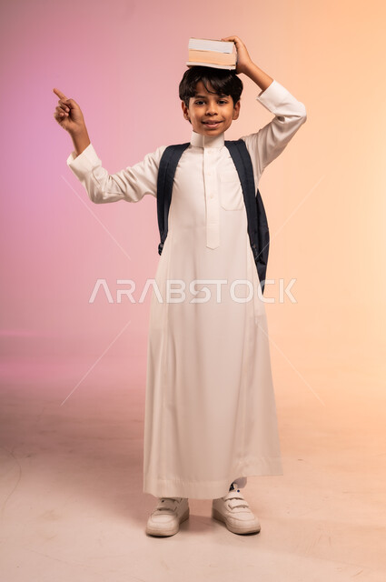  النظر للكاميرا بإيماءات السرور، الاستعداد للذهاب إلى المدرسة، بورتريه لطالب عربي خليجي سعودي يرتدي الثوب التقليدي يحمل حقيبة الظهر المدرسية يضع الكتب العلمية فوق رأسه، موسم العودة للمدارس في المملكة العربية السعودية، صورة الجسم بالطول الكامل، خلفية ملونة