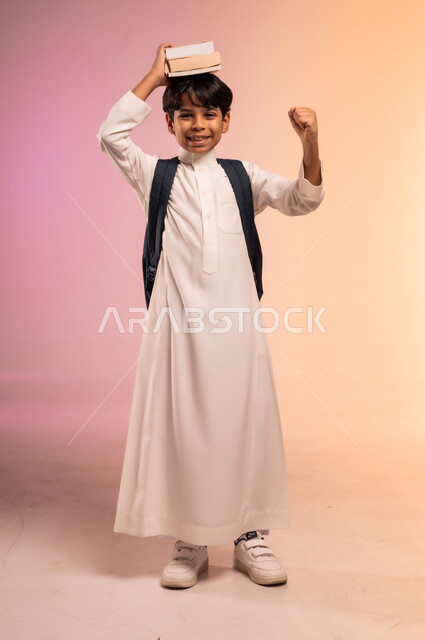  النظر للكاميرا بإيماءات السرور، الاستعداد للذهاب إلى المدرسة، بورتريه لطالب عربي خليجي سعودي يرتدي الثوب التقليدي يحمل حقيبة الظهر المدرسية يضع الكتب العلمية فوق رأسه، موسم العودة للمدارس في المملكة العربية السعودية، صورة الجسم بالطول الكامل، خلفية ملونة