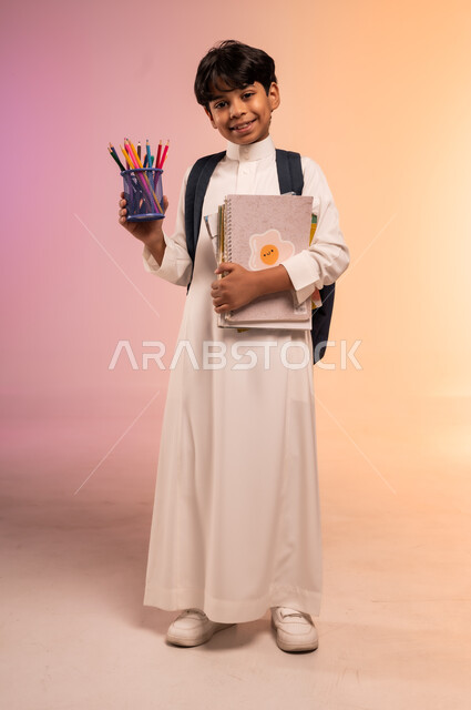 عرض القرطاسية المدرسية الجديدة، الاستعداد للعام الدراسي بإيماءات السرور والسعادة، بورتريه لطالب عربي خليجي سعودي يرتدي الثوب التقليدي يحمل حقيبة الظهر السوداء و يمسك بمجوعة من الأقلام والدفاتر الأنيقة، صورة الجسم بالطول الكامل، خلفية ملونة
