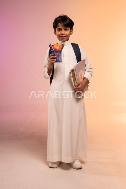 عرض القرطاسية المدرسية الجديدة، الاستعداد للعام الدراسي بإيماءات السرور والسعادة، بورتريه لطالب عربي خليجي سعودي يرتدي الثوب التقليدي يحمل حقيبة الظهر السوداء و يمسك بمجوعة من الأقلام والدفاتر الأنيقة، صورة الجسم بالطول الكامل، خلفية ملونة