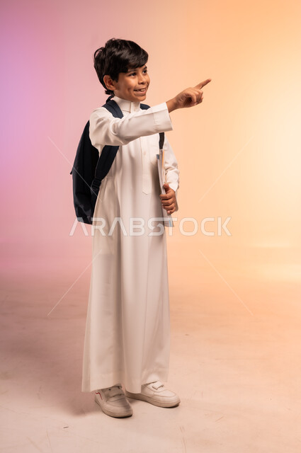 الإشارة باليد إلى شيء ما بعيد، موسم العودة إلى المدارس، الجد والاجتهاد للوصول إلى النجاح والتفوق، بورتريه لطالب عربي خليجي سعودي يرتدي الثوب التقليدي يحمل حقيبة الظهر المدرسية ينظر للأعلى بإيماءات التركيز والاندماج، الجسم بالطول الكامل، خلفية ملونة