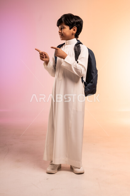  الوقوف بظهر مشدود مستقيم بثقة بالنفس، الابتهاج بموسم العودة إلى المدارس، بورتريه لطالب عربي خليجي سعودي يرتدي الثوب التقليدي ويحمل حقيبة الظهر المدرسية يرقع يديه ويششر لمكان ما، النظر للكاميرا بتعابير السرور والسعادة، الجسم بالطول الكامل، خلفية ملونة