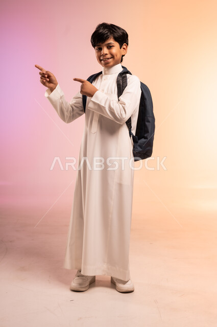  الوقوف بظهر مشدود مستقيم بثقة بالنفس، الابتهاج بموسم العودة إلى المدارس، بورتريه لطالب عربي خليجي سعودي يرتدي الثوب التقليدي ويحمل حقيبة الظهر المدرسية يرقع يديه ويششر لمكان ما، النظر للكاميرا بتعابير السرور والسعادة، الجسم بالطول الكامل، خلفية ملونة