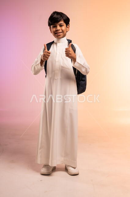 إشارات يد تدل على الموافقة والجودة، الوقوف بظهر مشدود واستقامة وثقة بالنفس، موسم العودة إلى المدارس، بورتريه لطالب عربي خليجي سعودي يرتدي الثوب التقليدي ويحمل حقيبة الظهر المدرسية ينظر للكاميرا بتعابير الابتهاج والفرح، الجسم بالطول الكامل، خلفية ملونة