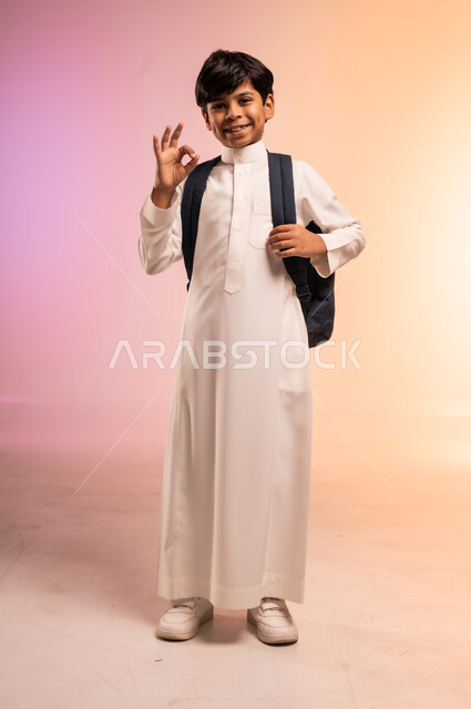إشارات يد تدل على الموافقة والجودة، الوقوف بظهر مشدود واستقامة وثقة بالنفس، موسم العودة إلى المدارس، بورتريه لطالب عربي خليجي سعودي يرتدي الثوب التقليدي ويحمل حقيبة الظهر المدرسية ينظر للكاميرا بتعابير الابتهاج والفرح، الجسم بالطول الكامل، خلفية ملونة