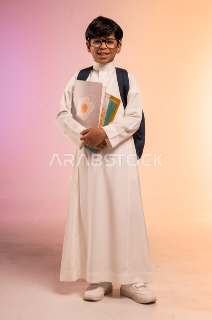 التطلع نحو النجاح والتفوق، موسم العودة إلى المدارس، بورتريه لطالب عربي خليجي سعودي يرتدي الثوب التقليدي ويضع نظارة طبية يحمل حقيبة الظهر المدرسية، الإمساك بالكتب والمناهج التعليمية والنظر للكاميرا بإيماءات السرور، الجسم بالطول الكامل، خلفية ملونة
