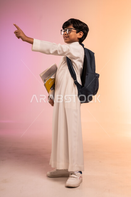 النظر إلى الأعلى بإيماءات التركيز والاندماج،موسم العودة إلى المدارس، الجد والاجتهاد للوصول إلى النجاح والتفوق، بورتريه لطالب عربي خليجي سعودي يرتدي الثوب التقليدي ويضع نظارة طبية يحمل حقيبة الظهر المدرسية ويشيرإلى شيء ما بعيد، الجسم بالطول الكامل، خلفية ملونة
