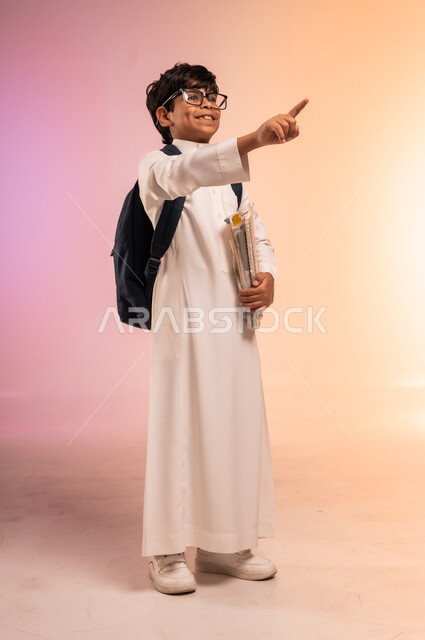 النظر إلى الأعلى بإيماءات التركيز والاندماج،موسم العودة إلى المدارس، الجد والاجتهاد للوصول إلى النجاح والتفوق، بورتريه لطالب عربي خليجي سعودي يرتدي الثوب التقليدي ويضع نظارة طبية يحمل حقيبة الظهر المدرسية ويشيرإلى شيء ما بعيد، الجسم بالطول الكامل، خلفية ملونة