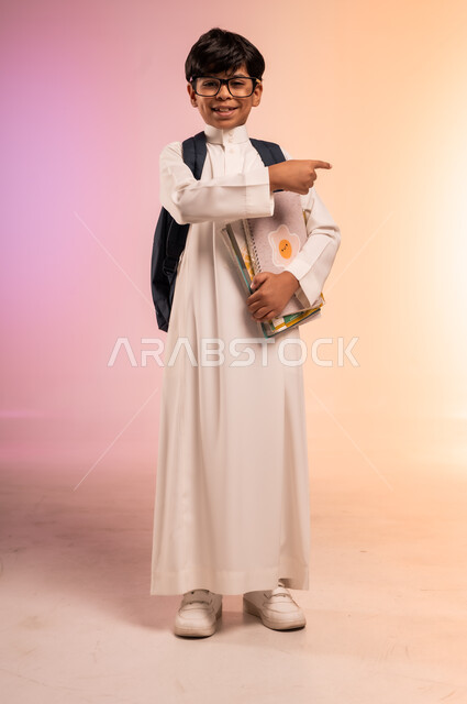 إيماءات السعادة والسرور، موسم العودة إلى المدارس، الجد والاجتهاد للوصول إلى النجاح والتفوق، بورتريه لطالب عربي خليجي سعودي يرتدي الثوب التقليدي ويضع نظارة طبية يحمل حقيبة الظهر المدرسية ويمسك بالكتب والمناهج التعليمية، الجسم بالطول الكامل، خلفية ملونة