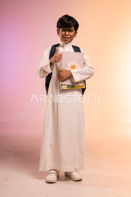 Gestures of happiness and pleasure, back to school season, hard work and diligence to achieve success and excellence, portrait of a Saudi Arabian Gulf student wearing traditional dress and glasses carrying a school backpack and holding books and educational curricula, full body, colored background