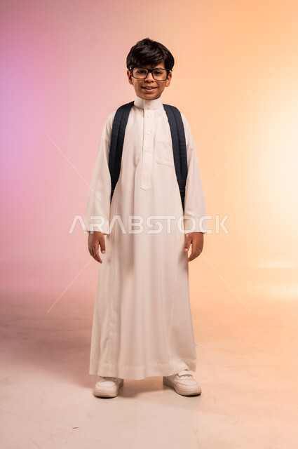 الوقوف بظهر مشدود واستقامة، موسم العودة إلى المدارس، بورتريه لطالب عربي خليجي سعودي يرتدي الثوب التقليدي يضع نظارة طبية ويحمل حقيبة الظهر المدرسية ينظر للكاميرا بإيماءات السرور والسعادة، الجسم بالطول الكامل، خلفية ملونة