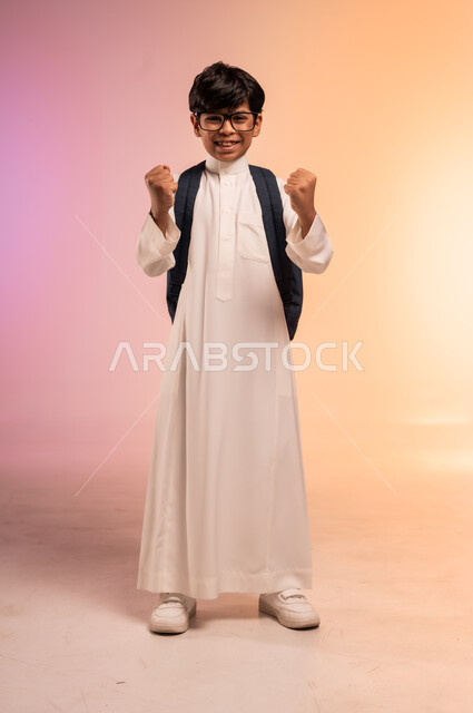 رفع اليدين بإيماءات الحماس والسعادة، الوقوف بظهر مشدود واستقامة، موسم العودة إلى المدارس، بورتريه لطالب عربي خليجي سعودي يرتدي الثوب التقليدي يضع نظارة طبية ويحمل حقيبة الظهر المدرسية ينظر للكاميرا بتعابير الابتهاج والفرح، الجسم بالطول الكامل، خلفية ملونة
