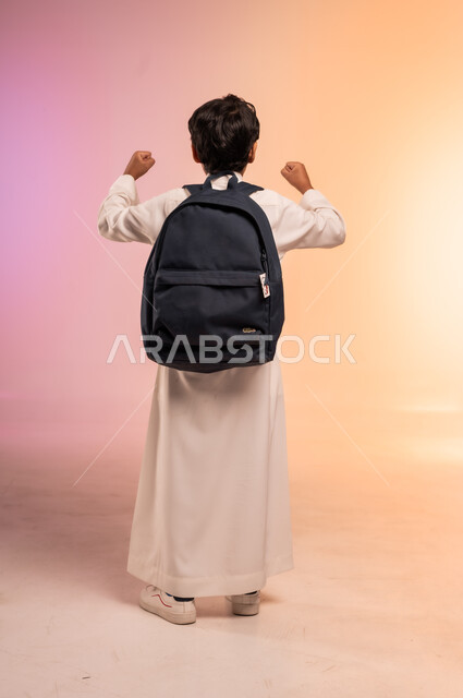 الاستعداد للعام الدراسي الجديد، موسم العودة إلى المدارس، بورتريه من الخلف لطالب عربي خليجي سعودي يرتدي الثوب التقليدي يحمل حقيبة الظهر المدرسية يرفع يديه في الهواء بإيماءات الحماس والابتهاج، صورة الجسم بالطول الكامل، خلفية ملونة