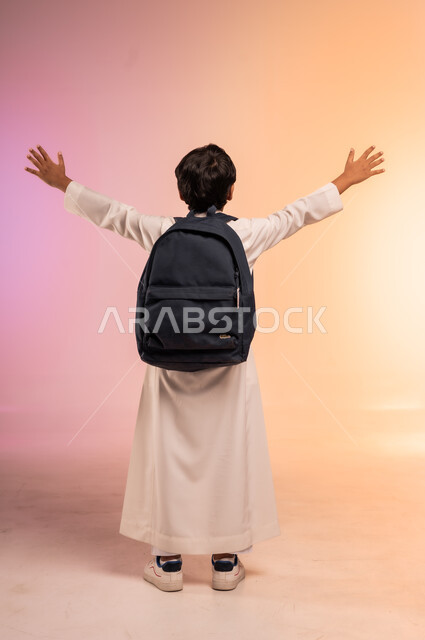 الاستعداد للعام الدراسي الجديد، موسم العودة إلى المدارس، بورتريه من الخلف لطالب عربي خليجي سعودي يرتدي الثوب التقليدي يحمل حقيبة الظهر المدرسية يرفع يديه في الهواء بإيماءات الحماس والابتهاج، صورة الجسم بالطول الكامل، خلفية ملونة