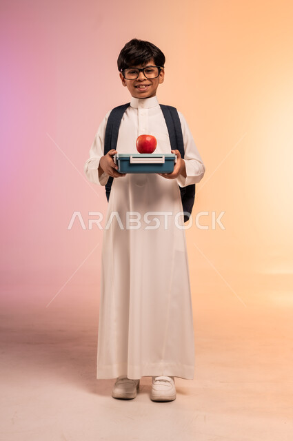السناكات الصحية المغذية، موسم العودة إلى المدارس، بورتريه لطالب عربي خليجي سعودي يرتدي الثوب التقليدي يضع نظارة طبية ويحمل حقيبة الظهر المدرسية، الإمساك بعلبة الطعام والنظر للكاميرا بإيماءات السرور، الجسم بالطول الكامل، خلفية ملونة