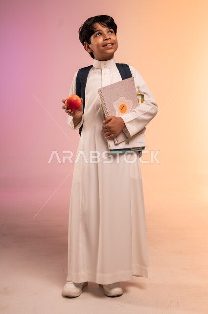 النظر للأعلى بإيماءات الأمل والحلم بالتفوق، موسم العودة للمدارس في السعودية، الغذاء والوجبات الصحية خلال الدوام، بورتريه لطالب عربي خليجي سعودي يرتدي الثوب التقليدي وحقيبة الظهر المدرسية يقف ويحمل الكتب ويمسك تفاحة حمراء، الجسم بالطول الكامل، خلفية ملونة