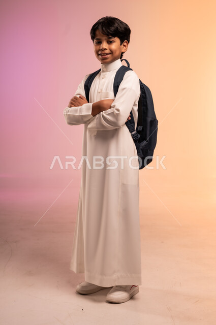 السعي نحو تحقيق حلم النجاح والتفوق، بورتريه لطالب عربي خليجي سعودي يرتدي الثوب التقليدي يحمل حقيبة الظهر المدرسية يقف بيدين متقاطعتين وينظر للكاميرا بإيماءات الثقة والبشاشة، موسم العودة للمدارس في المملكة العربية السعودية، الجسم بالطول الكامل، خلفية ملونة