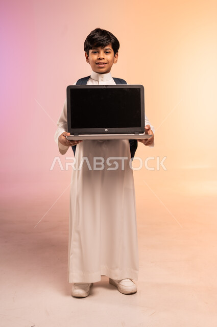 دمج التقنيات الحديثة بالتعليم، الاستعداد للعام الدراسي الجديد بإيماءات السرور، موسم العودة إلى المدارس، بورتريه لطالب عربي خليجي سعودي يرتدي الثوب التقليدي يحمل حقيبة الظهر المدرسية يمسك الحاسوب المحمول بيديه، صورة الجسم بالطول الكامل، خلفية ملونة