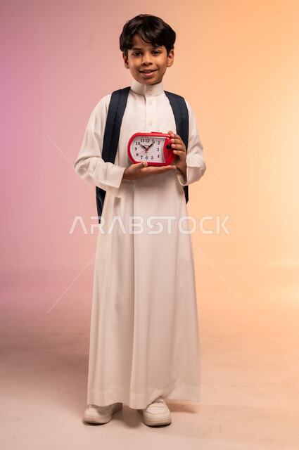 أهمية الاستيقاظ باكرا للذهاب إلى المدرسة، بورتريه لطالب عربي خليجي سعودي يرتدي الثوب التقليدي يحمل حقيبة الظهر المدرسية يمسك ساعة المنبه بيده وينظر للكاميرا بإيماءات البشاشة، موسم العودة للمدارس في المملكة العربية السعودية، الجسم بالطول الكامل، خلفية ملونة