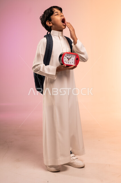 الإمساك بساعة المنبه و أهمية الاستيقاظ باكرا للذهاب إلى المدرسة، بورتريه لطالب عربي خليجي سعودي يرتدي الثوب التقليدي يحمل حقيبة الظهر المدرسية يشعر بالنعاس ويقوم بالتثاؤب، موسم العودة للمدارس في المملكة العربية السعودية، الجسم بالطول الكامل، خلفية ملونة