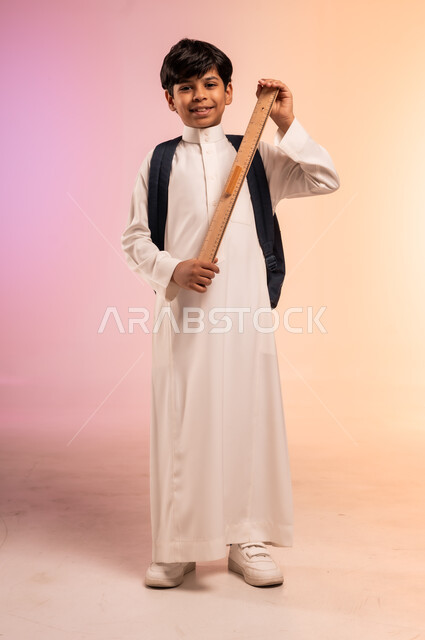 الإمساك بالمسطرة الخشبية، الوقوف بظهر مشدود مستقيم وثقة بالنفس، موسم العودة إلى المدارس والقرطاسية الجديدة، بورتريه لطالب عربي خليجي سعودي يرتدي الثوب التقليدي ويحمل حقيبة الظهر المدرسية ينظر للكاميرا بتعابير الابتهاج والفرح، الجسم بالطول الكامل، خلفية ملونة