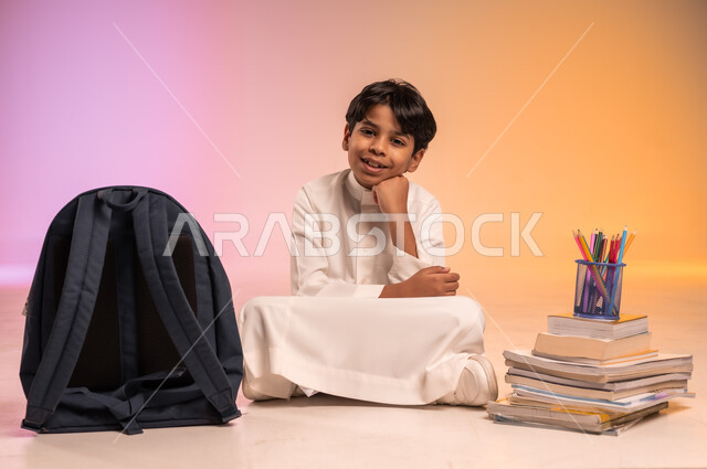 النظر للكاميرا بإيماءات السعادة، تجهيز المستلزمات المدرسية لبدء موسم دراسي جديد، الاستعداد لافتتاح المدارس في المملكة العربية السعودية، بورتريه لولد عربي خليجي سعودي يرتدي الثوب التقليدي يجلس بين كومة من الكتب والقرطاسية وأمامه ساعة المنبه، خلفية ملونة