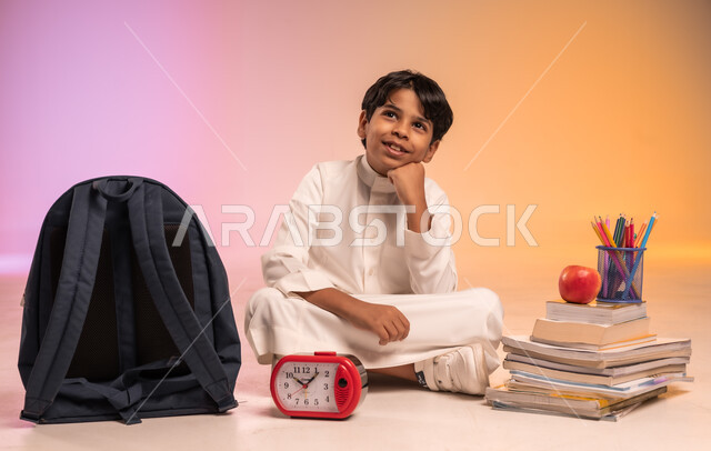 تجهيز المستلزمات المدرسية لبدء موسم دراسي جديد، الاستعداد لافتتاح المدارس في المملكة العربية السعودية، بورتريه لولد عربي خليجي سعودي يرتدي الثوب التقليدي يجلس بين كومة من الكتب والقرطاسية وأمامه ساعة المنبه، النظر للأعلى والحلم بالنجاح والتفوق، خلفية ملونة