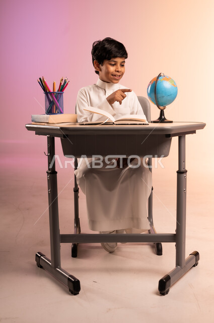 حل الواجبات ومراجعة الدروس، الجد والاجتهاد للوصول إلى النجاح والتفوق، إيماءات السعادة والسرور بالعودة إلى المدارس، بورتريه لطالب عربي خليجي سعودي مبتسم يرتدي الثوب التقليدي يجلس في مقعده يقوم بالدراسة والكتابة، قرطاسية مدرسية ومجسم للكرة الأرضية، خلفية ملونة
