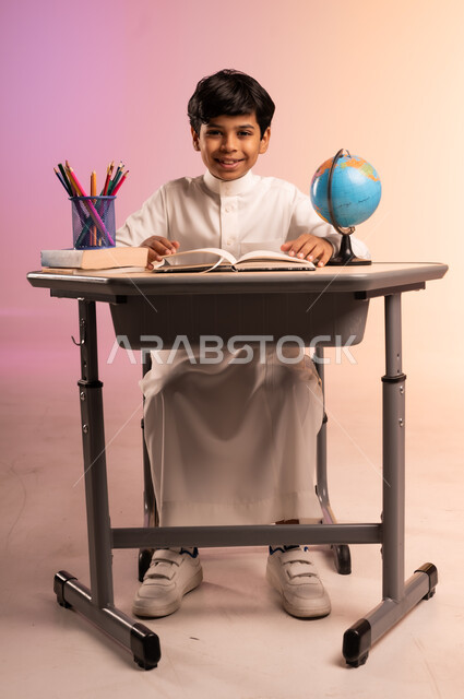 حل الواجبات ومراجعة الدروس، الجد والاجتهاد للوصول إلى النجاح والتفوق، إيماءات السعادة والسرور بالعودة إلى المدارس، بورتريه لطالب عربي خليجي سعودي مبتسم يرتدي الثوب التقليدي يجلس في مقعده يقوم بالدراسة والكتابة، قرطاسية مدرسية ومجسم للكرة الأرضية، خلفية ملونة