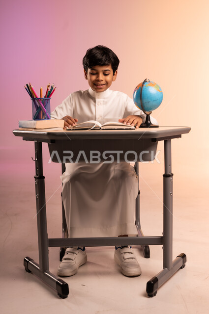 حل الواجبات ومراجعة الدروس، الجد والاجتهاد للوصول إلى النجاح والتفوق، إيماءات السعادة والسرور بالعودة إلى المدارس، بورتريه لطالب عربي خليجي سعودي مبتسم يرتدي الثوب التقليدي يجلس في مقعده يقوم بالدراسة والكتابة، قرطاسية مدرسية ومجسم للكرة الأرضية، خلفية ملونة