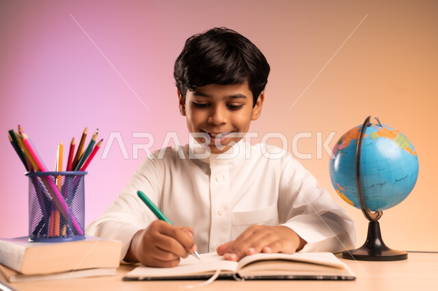 حل الواجبات ومراجعة الدروس، الجد والاجتهاد للوصول إلى النجاح والتفوق، إيماءات السعادة والسرور بالعودة إلى المدارس، بورتريه لطالب عربي خليجي سعودي مبتسم يرتدي الثوب التقليدي يجلس في مقعده يقوم بالدراسة والكتابة، قرطاسية مدرسية ومجسم للكرة الأرضية، خلفية ملونة