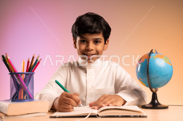 حل الواجبات ومراجعة الدروس، الجد والاجتهاد للوصول إلى النجاح والتفوق، إيماءات السعادة والسرور بالعودة إلى المدارس، بورتريه لطالب عربي خليجي سعودي مبتسم يرتدي الثوب التقليدي يجلس في مقعده يقوم بالدراسة والكتابة، قرطاسية مدرسية ومجسم للكرة الأرضية، خلفية ملونة