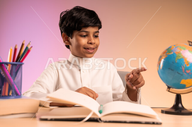 النظر إلى مجسم للكرة الأرضية بإيماءات الاندماج والتركيز، حل الواجبات ومراجعة الدروس، الجد والاجتهاد للوصول إلى النجاح والتفوق، العودة إلى المدارس، بورتريه لطالب عربي خليجي سعودي مبتسم يرتدي الثوب التقليدي يجلس في مقعده، التفكير حول شيء ما، خلفية ملونة