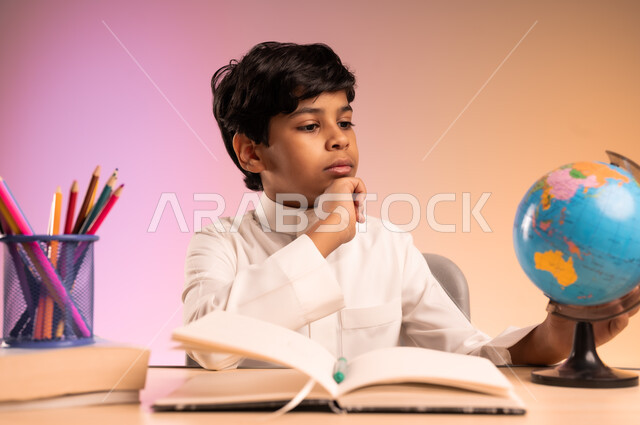 Looking at a globe with gestures of integration and concentration, doing homework and reviewing lessons, seriousness and diligence to achieve success and excellence, back to school, portrait of a smiling Saudi Arabian Gulf student wearing traditional dress sitting in his seat, thinking about something, colorful background