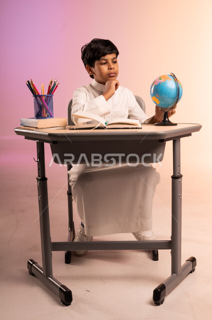 Looking at a globe with gestures of integration and concentration, doing homework and reviewing lessons, seriousness and diligence to achieve success and excellence, back to school, portrait of a smiling Saudi Arabian Gulf student wearing traditional dress sitting in his seat, thinking about something, colorful background