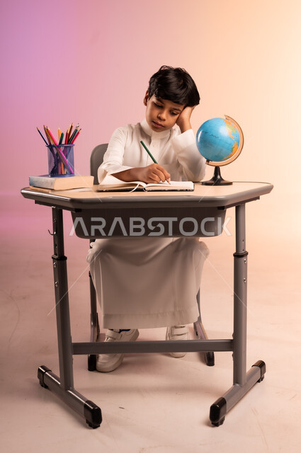 Gestures of integration and concentration in solving homework and reviewing lessons, seriousness and diligence to achieve success and excellence, back to school, portrait of a smiling Saudi Arabian Gulf student wearing traditional dress sitting in his seat writing, school stationery and a globe, colorful background