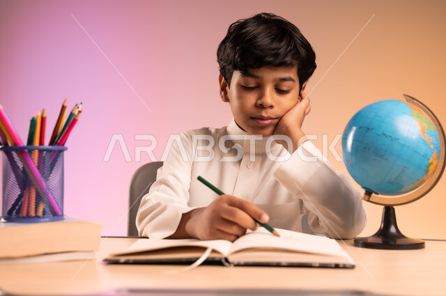 إيماءات الاندماج والتركيز في حل الواجبات ومراجعة الدروس، الجد والاجتهاد للوصول إلى النجاح والتفوق، العودة إلى المدارس، بورتريه لطالب عربي خليجي سعودي مبتسم يرتدي الثوب التقليدي يجلس في مقعده يقوم بالكتابة، قرطاسية مدرسية ومجسم للكرة الأرضية، خلفية ملونة