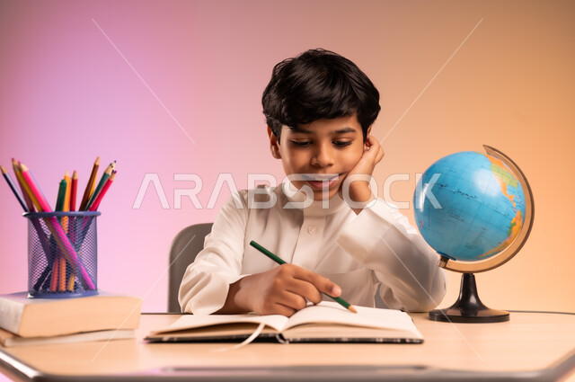 إيماءات الاندماج والتركيز في حل الواجبات ومراجعة الدروس، الجد والاجتهاد للوصول إلى النجاح والتفوق، العودة إلى المدارس، بورتريه لطالب عربي خليجي سعودي مبتسم يرتدي الثوب التقليدي يجلس في مقعده يقوم بالكتابة، قرطاسية مدرسية ومجسم للكرة الأرضية، خلفية ملونة