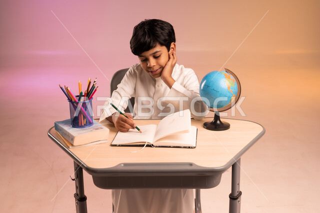 Gestures of integration and concentration in solving homework and reviewing lessons, seriousness and diligence to achieve success and excellence, back to school, portrait of a smiling Saudi Arabian Gulf student wearing traditional dress sitting in his seat writing, school stationery and a globe, colorful background