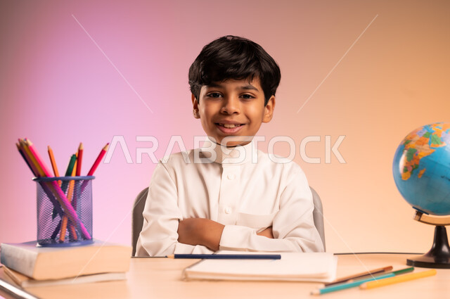 الجد والاجتهاد للوصول إلى النجاح والتفوق، إيماءات السعادة والسرور بالعودة إلى المدارس، بورتريه لطالب عربي خليجي سعودي يرتدي الثوب التقليدي يجلس في مقعده ينظر للكاميرا ويبتسم، قرطاسية مدرسية ومجسم للكرة الأرضية، خلفية ملونة
