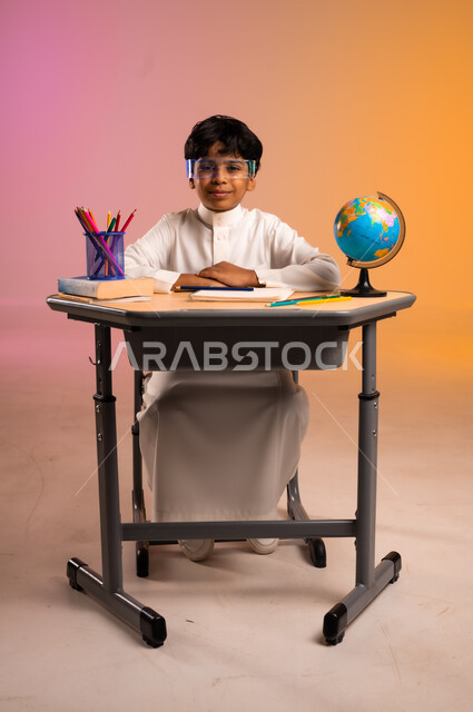 ارتداء النظارات المخبرية والتفاعل مع الدروس، الجد والاجتهاد للوصول إلى النجاح والتفوق، إيماءات السعادة والسرور بالعودة إلى المدارس، بورتريه لطالب عربي خليجي سعودي مبتسم يرتدي الثوب التقليدي يجلس في مقعده، قرطاسية مدرسية ومجسم للكرة الأرضية، خلفية ملونة