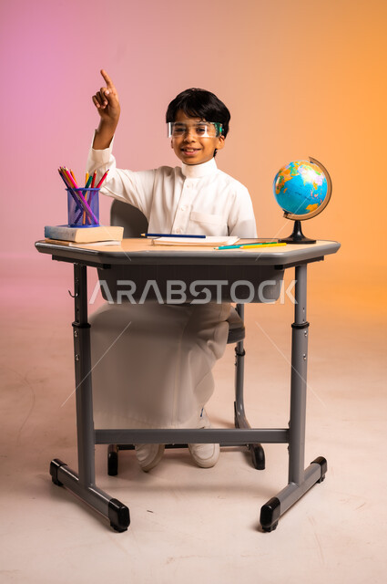 ارتداء النظارات المخبرية والتفاعل مع الدروس، الجد والاجتهاد للوصول إلى النجاح والتفوق، إيماءات السعادة والسرور بالعودة إلى المدارس، بورتريه لطالب عربي خليجي سعودي مبتسم يرتدي الثوب التقليدي يجلس في مقعده، قرطاسية مدرسية ومجسم للكرة الأرضية، خلفية ملونة