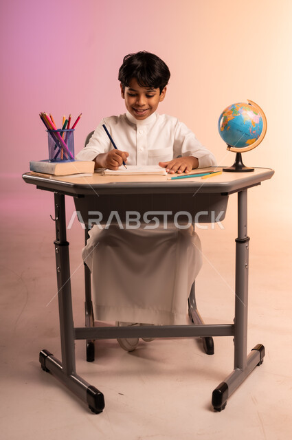 إيماءات السعادة والسرور بالعودة إلى المدارس، الجد والاجتهاد للوصول إلى النجاح والتفوق، بورتريه لطالب عربي خليجي سعودي يرتدي الثوب التقليدي يجلس في مقعده يمسك القلم بيده ويقوم بالكتابة والدراسة، قرطاسية مدرسية ومجسم للكرة الأرضية، خلفية ملونة