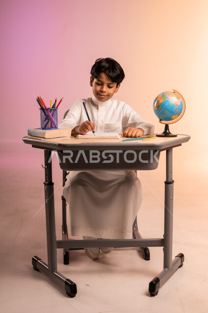 إيماءات السعادة والسرور بالعودة إلى المدارس، الجد والاجتهاد للوصول إلى النجاح والتفوق، بورتريه لطالب عربي خليجي سعودي يرتدي الثوب التقليدي يجلس في مقعده يمسك القلم بيده ويقوم بالكتابة والدراسة، قرطاسية مدرسية ومجسم للكرة الأرضية، خلفية ملونة