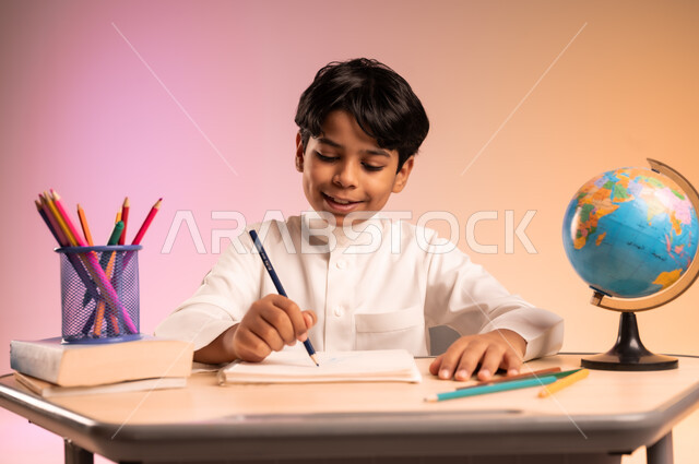 إيماءات السعادة والسرور بالعودة إلى المدارس، الجد والاجتهاد للوصول إلى النجاح والتفوق، بورتريه لطالب عربي خليجي سعودي يرتدي الثوب التقليدي يجلس في مقعده يمسك القلم بيده ويقوم بالكتابة والدراسة، قرطاسية مدرسية ومجسم للكرة الأرضية، خلفية ملونة
