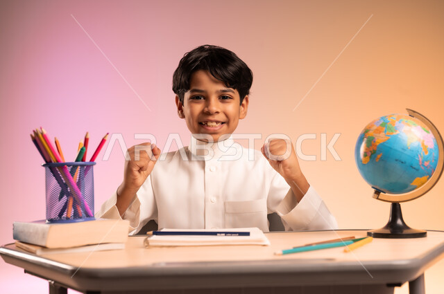 إيماءات السعادة والسرور بالعودة إلى المدارس، الجد والاجتهاد للوصول إلى النجاح والتفوق، بورتريه لطالب عربي خليجي سعودي يرتدي الثوب التقليدي يجلس في مقعده يرفع يديه وينظر للكاميرا بحماس، قرطاسية مدرسية ومجسم للكرة الأرضية، خلفية ملونة