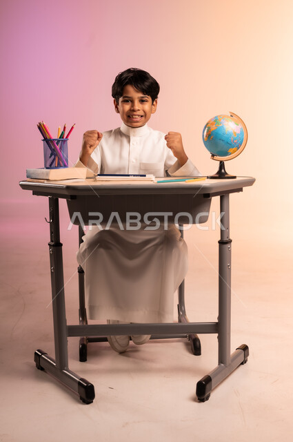 إيماءات السعادة والسرور بالعودة إلى المدارس، الجد والاجتهاد للوصول إلى النجاح والتفوق، بورتريه لطالب عربي خليجي سعودي يرتدي الثوب التقليدي يجلس في مقعده يرفع يديه وينظر للكاميرا بحماس، قرطاسية مدرسية ومجسم للكرة الأرضية، خلفية ملونة