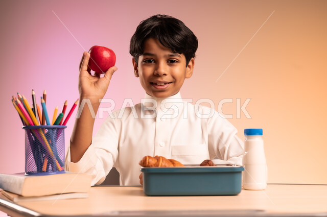 Lunch box with healthy snacks, back to school season in Saudi Arabia, gestures of happiness and joy, school stationery, pens and science books, portrait of a Saudi Arabian Gulf student wearing traditional dress sitting in his seat holding an apple from the lunch box in front of him, colorful background